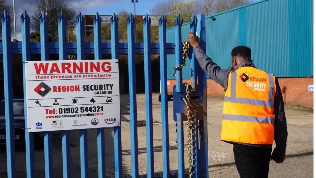 A Nuneaton security guard in an orange vest secures a blue gate with a chain, next to a warning sign indicating protection by Region Security Guarding