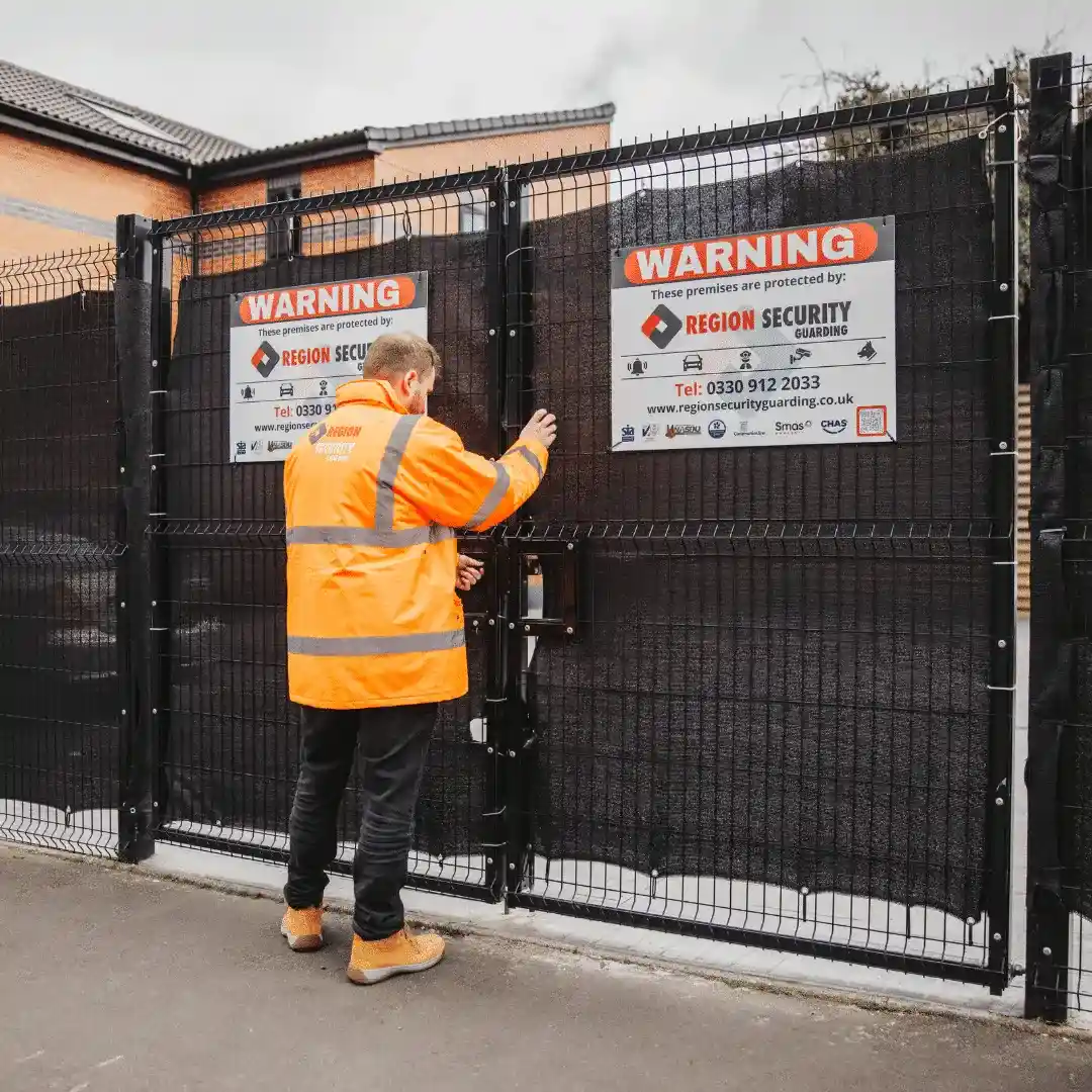 Security guard in an orange high-visibility jacket locking a gated fence with warning signs attached.